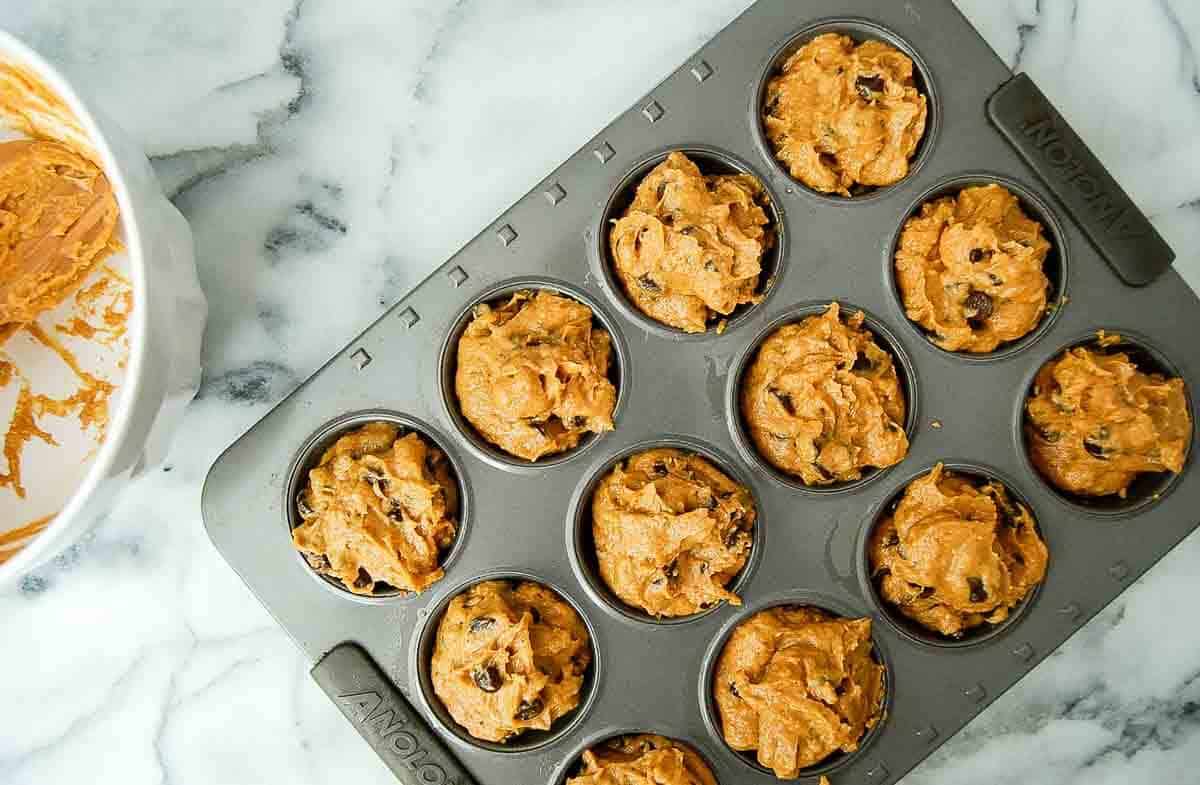 A muffin tin filled with unbaked pumpkin muffin batter containing chocolate chips sits on a marble countertop, with a mixing bowl and leftover batter visible nearby.