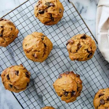 Pumpkin chocolate chip muffins cooling on a wire rack, placed on a marble surface with a white cloth partially visible on the side.