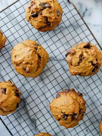 Pumpkin chocolate chip muffins cooling on a wire rack, placed on a marble surface with a white cloth partially visible on the side.