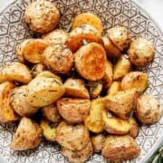 A bowl filled with roasted potato halves and quarters, garnished with fresh herbs, sits on a decorative plate with a geometric pattern. The potatoes appear crispy and seasoned.