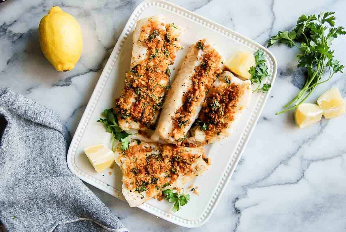 Four pieces of baked cod fish with a herb and breadcrumb topping served on a white plate, garnished with parsley and lemon wedges, with a lemon and parsley sprigs nearby on a marble surface.