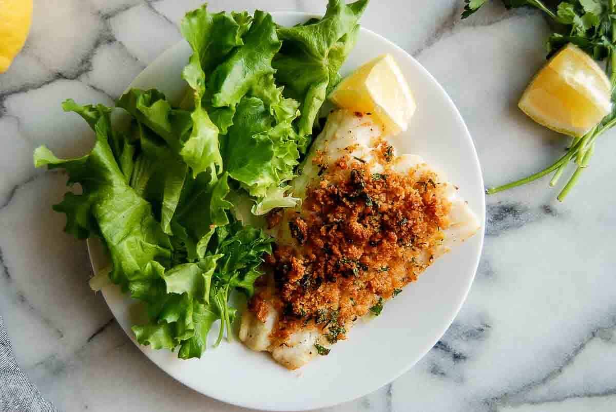A white plate with baked cod topped with golden breadcrumbs and herbs, served with fresh green lettuce leaves and lemon wedges, on a marble surface.