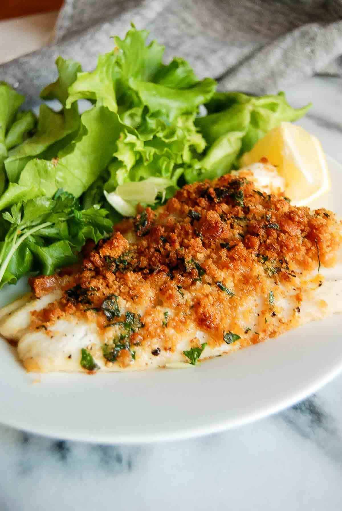 A baked cod fish fillet topped with golden, crispy breadcrumbs and herbs, served with fresh green lettuce, parsley, and a lemon wedge on a white plate.