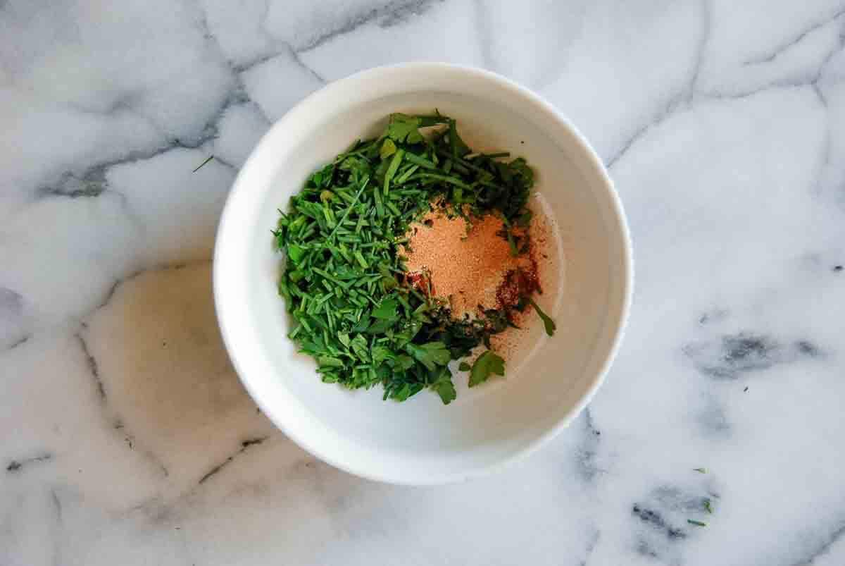 A white bowl on a marble surface contains chopped fresh green herbs and a small pile of reddish-brown and beige powdered spices.