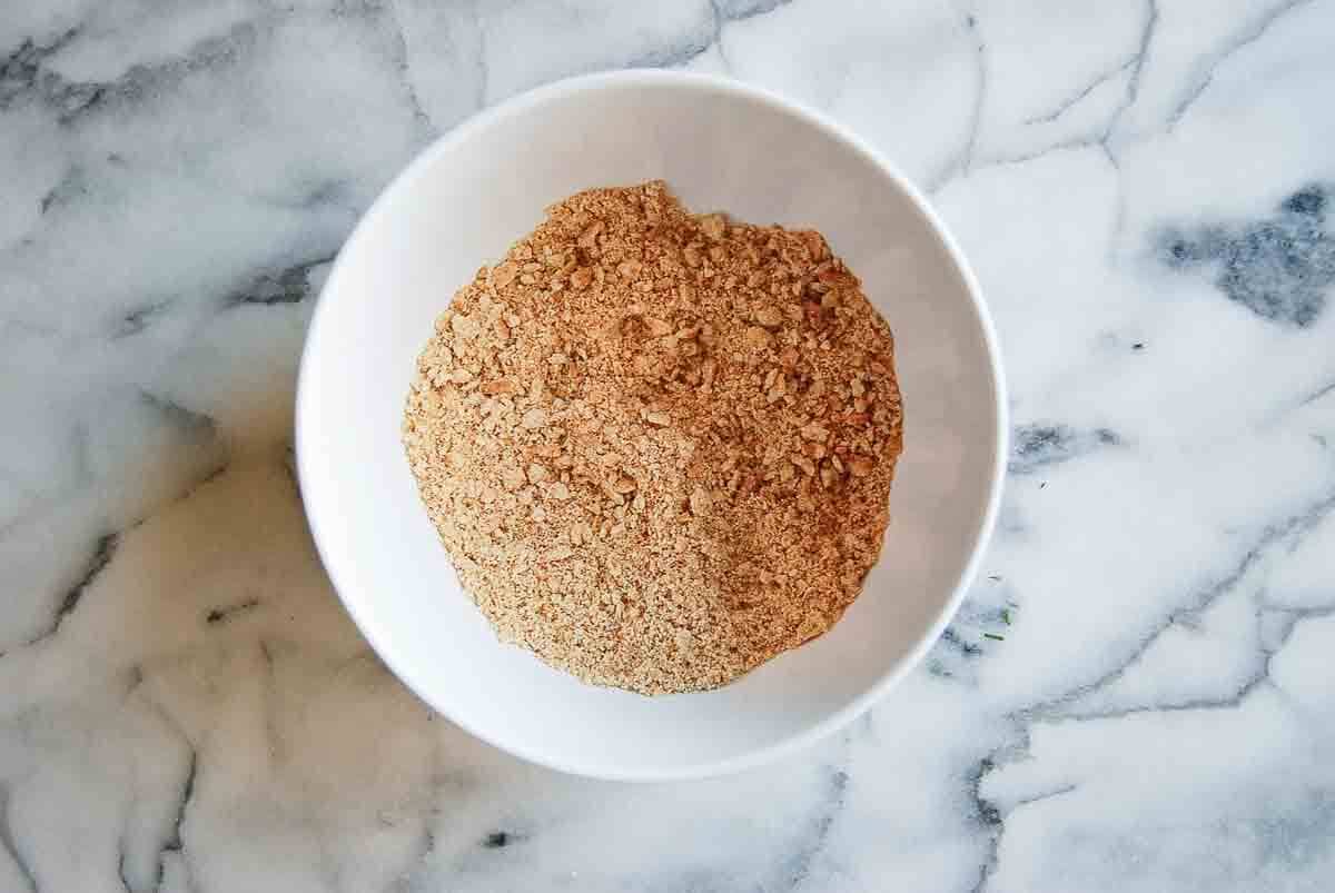 A white bowl filled with light brown, finely crushed graham cracker crumbs sits on a white marble surface.