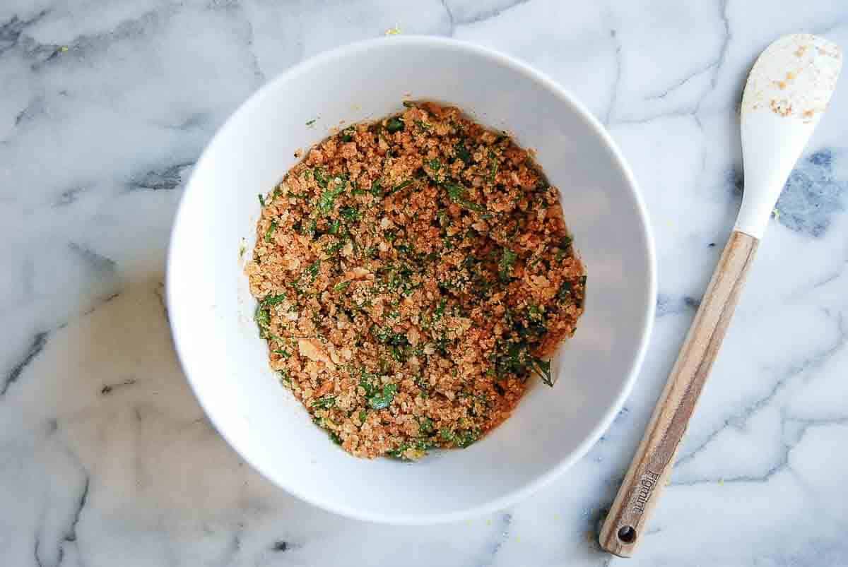 A white bowl filled with a breadcrumb and herb mixture sits on a marble surface next to a white spatula with a wooden handle.