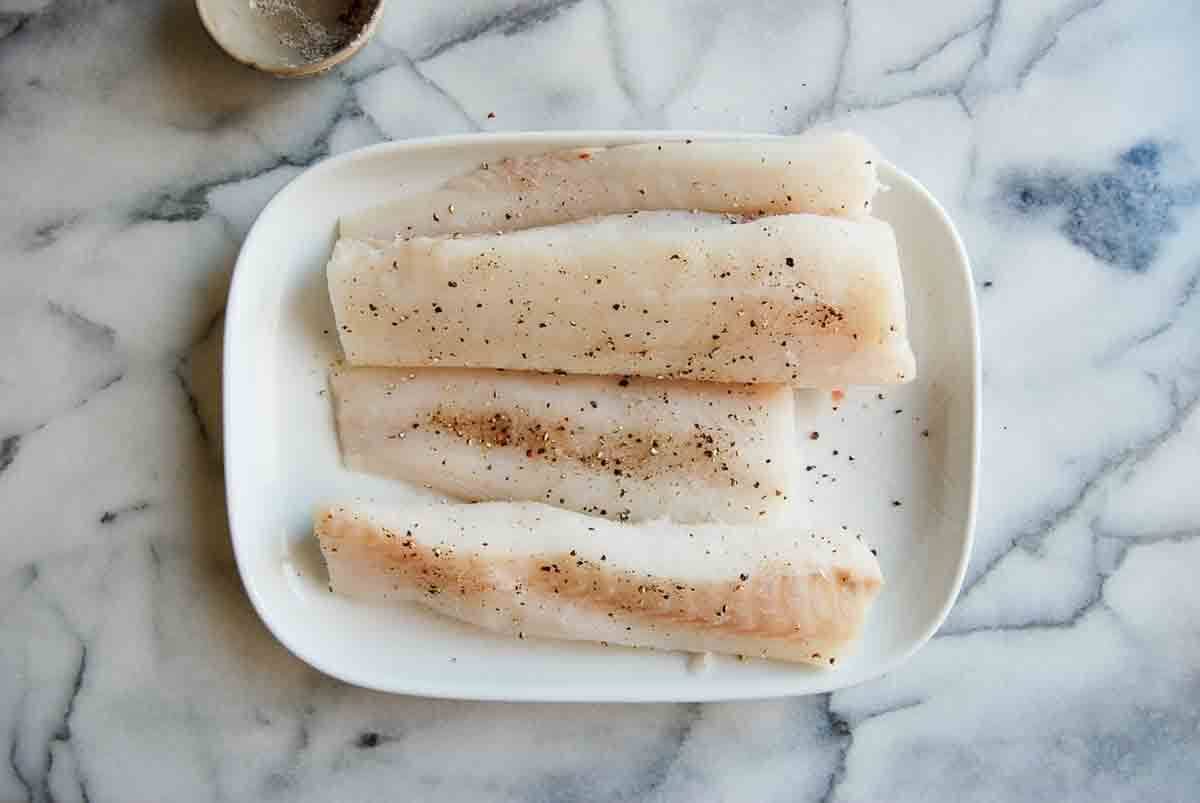 Four raw cod fillets sprinkled with salt and pepper are arranged on a white plate, set on a light marble surface. A small bowl with seasoning residue is visible in the upper left corner.