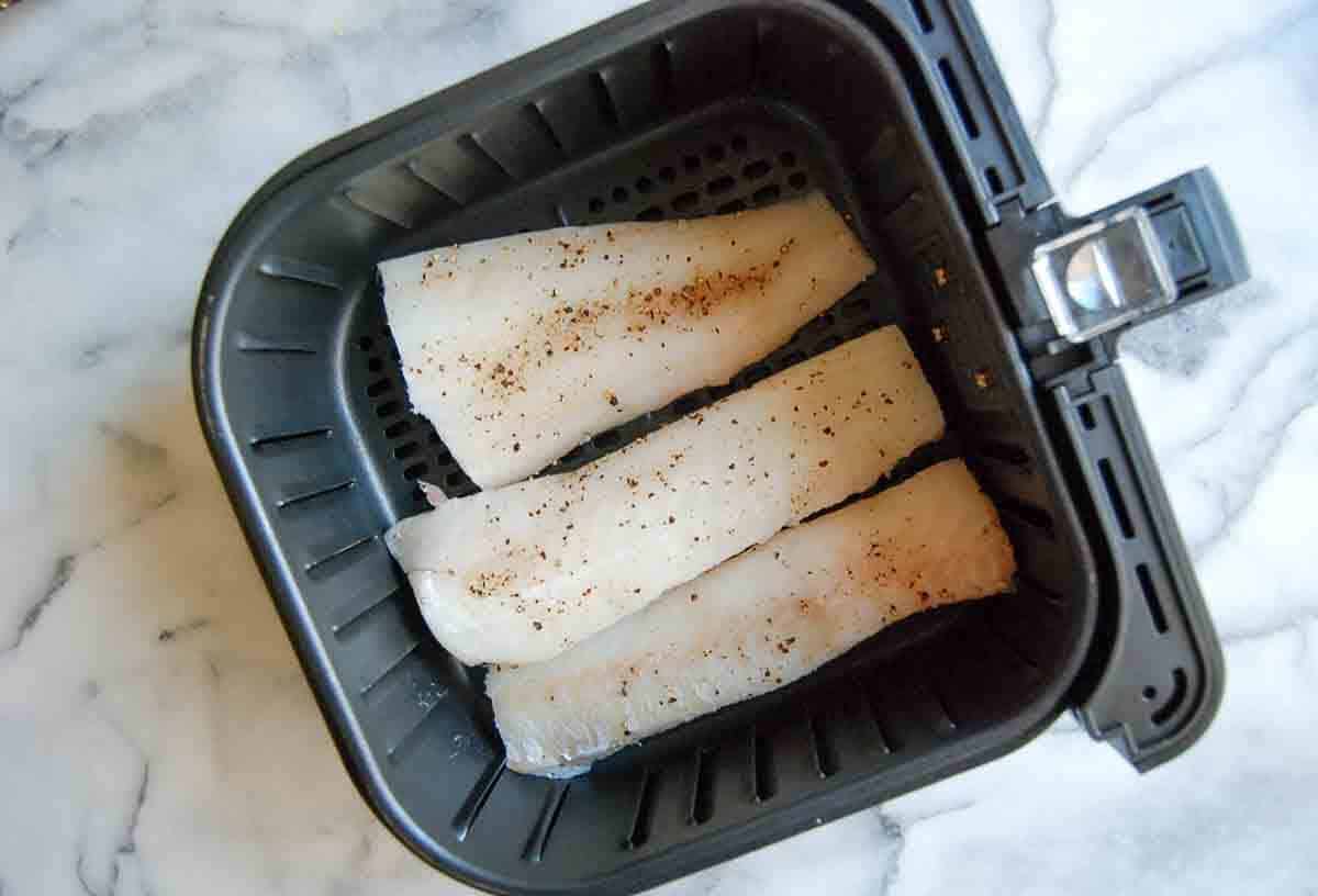 Three raw, seasoned cod fillets are placed inside the basket of an air fryer, ready to be cooked. The air fryer basket is black and sits on a white marble surface.