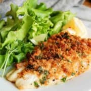 A plate with breaded, baked fish fillet garnished with chopped herbs, served alongside fresh green leafy lettuce and a lemon wedge.