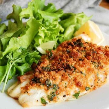 A plate with breaded, baked fish fillet garnished with chopped herbs, served alongside fresh green leafy lettuce and a lemon wedge.