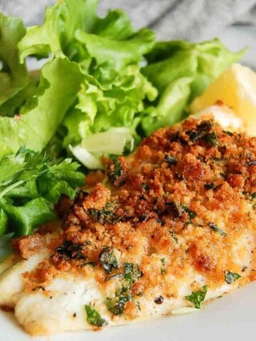 A plate with breaded, baked fish fillet garnished with chopped herbs, served alongside fresh green leafy lettuce and a lemon wedge.
