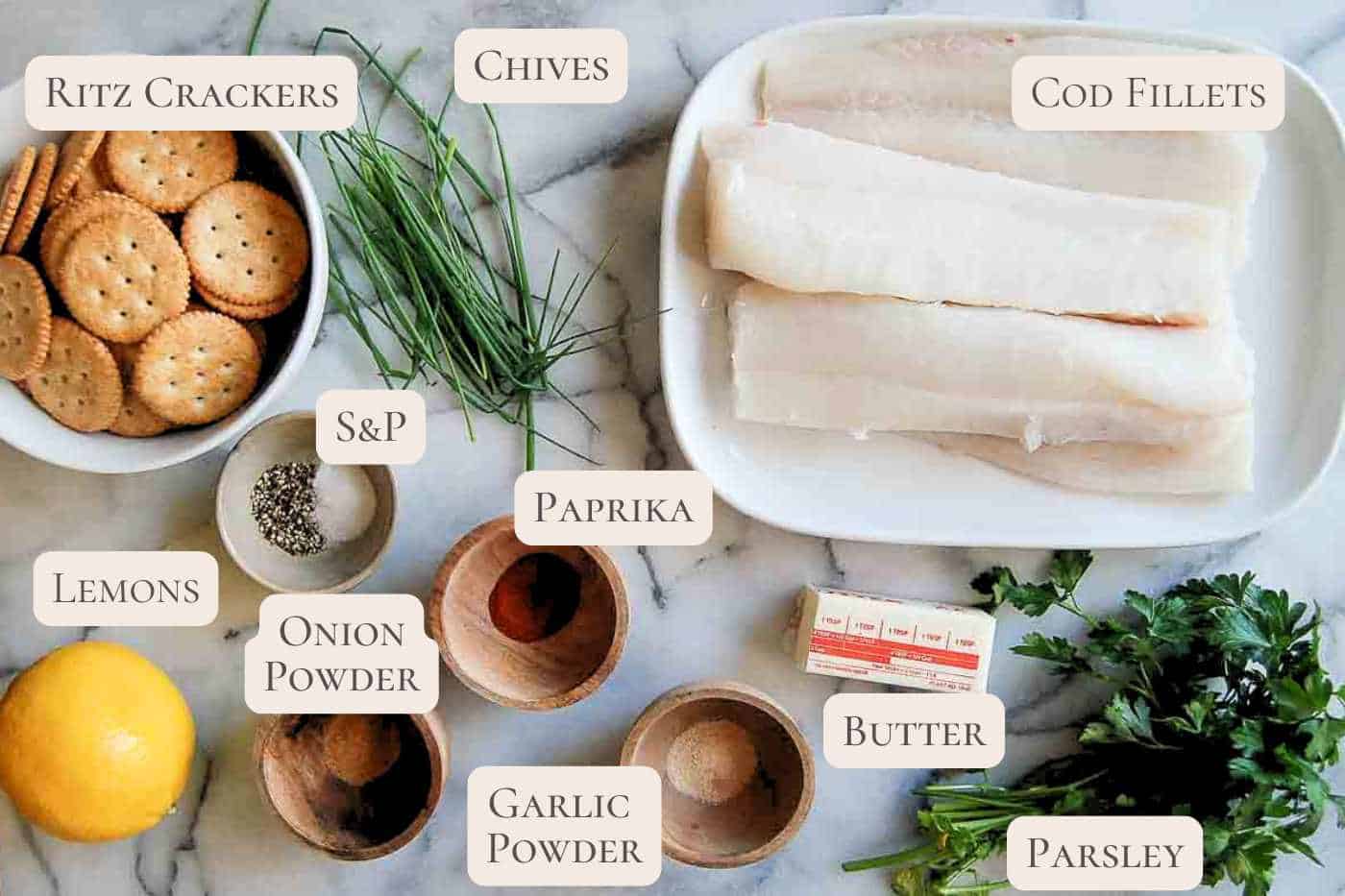 A marble surface displays ingredients for an air fryer cod recipe: cod fillets, Ritz crackers, chives, paprika, garlic powder, onion powder, salt and pepper, butter, parsley, and a lemon. Each ingredient is labeled with text.