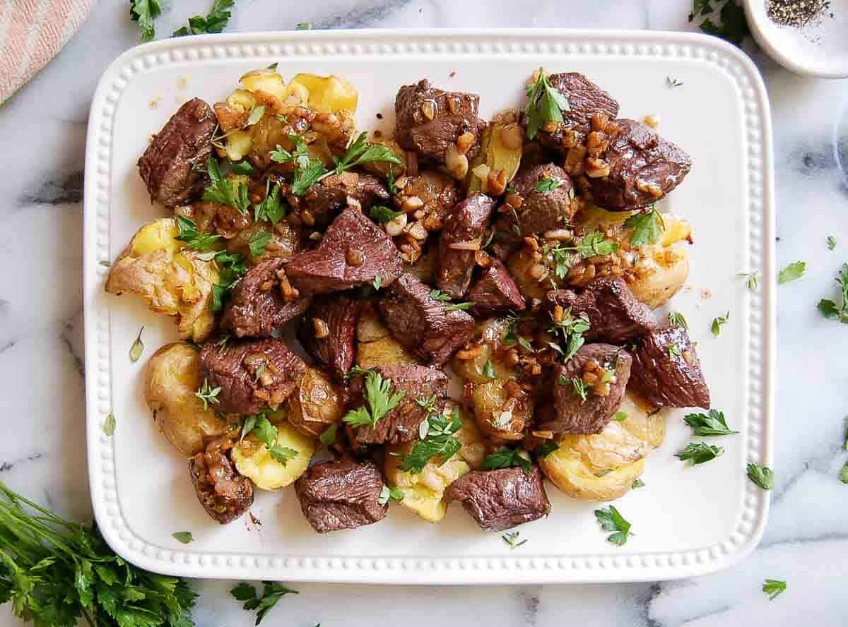 A white platter with browned steak bites and smashed potatoes, garnished with chopped parsley, garlic and herbs, rests on a marble surface. Scattered herbs and a small pepper dish are nearby.