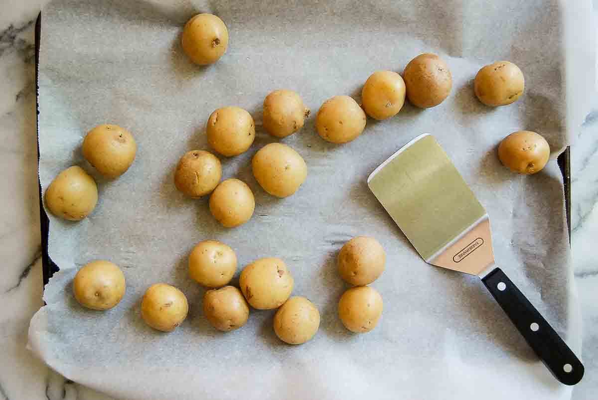 Small whole potatoes are scattered on a parchment-lined baking sheet next to a flat metal spatula with a black handle, all on a white marble surface.