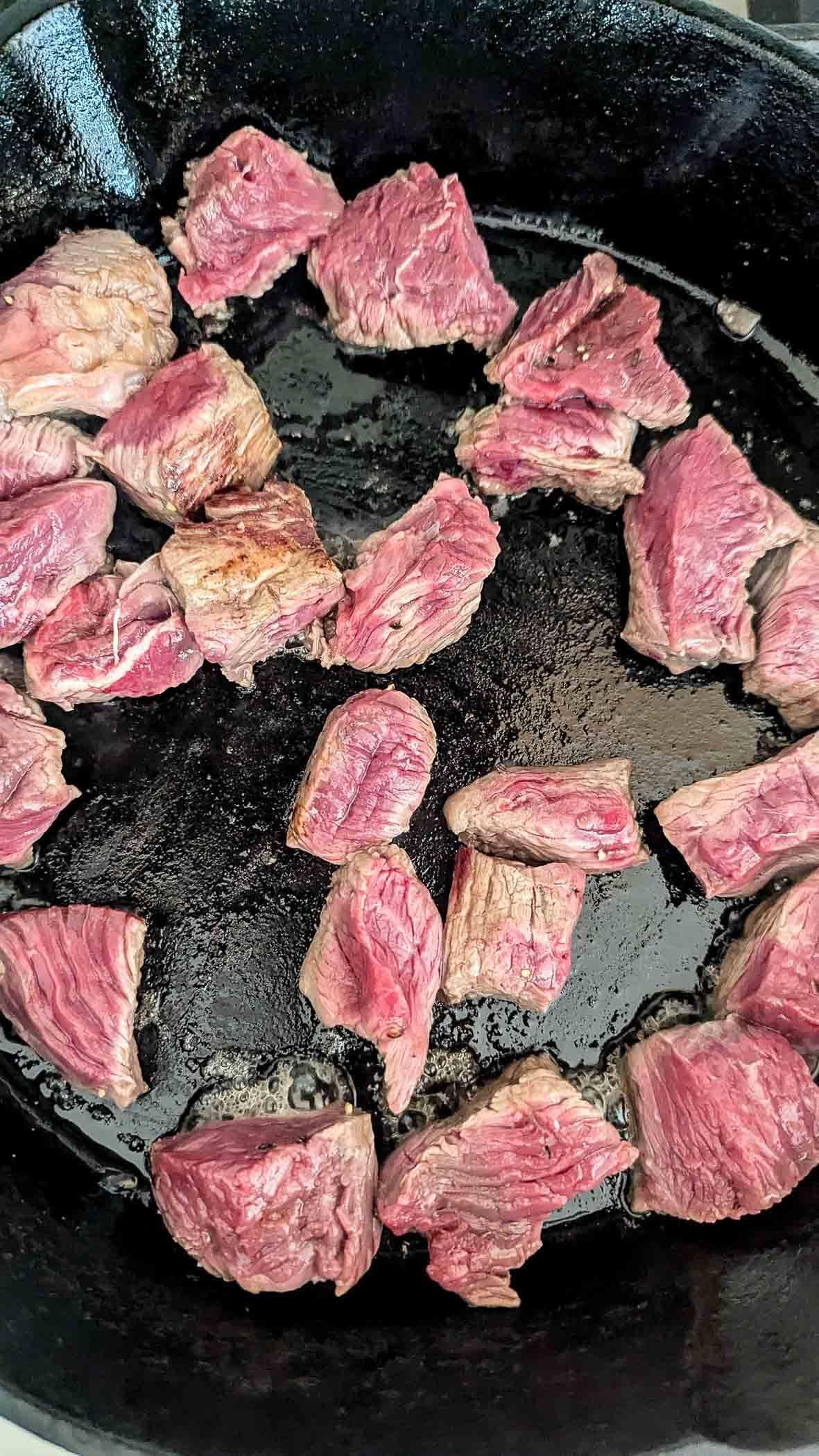 Chunks of beef searing in a black cast iron skillet, with some pieces browning on the outside while others remain red and raw in the center.