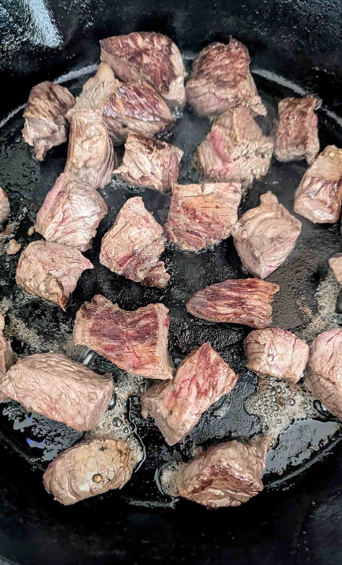 Chunks of beef searing and browning in a black cast iron skillet, with visible grill marks and sizzling juices around the meat pieces.
