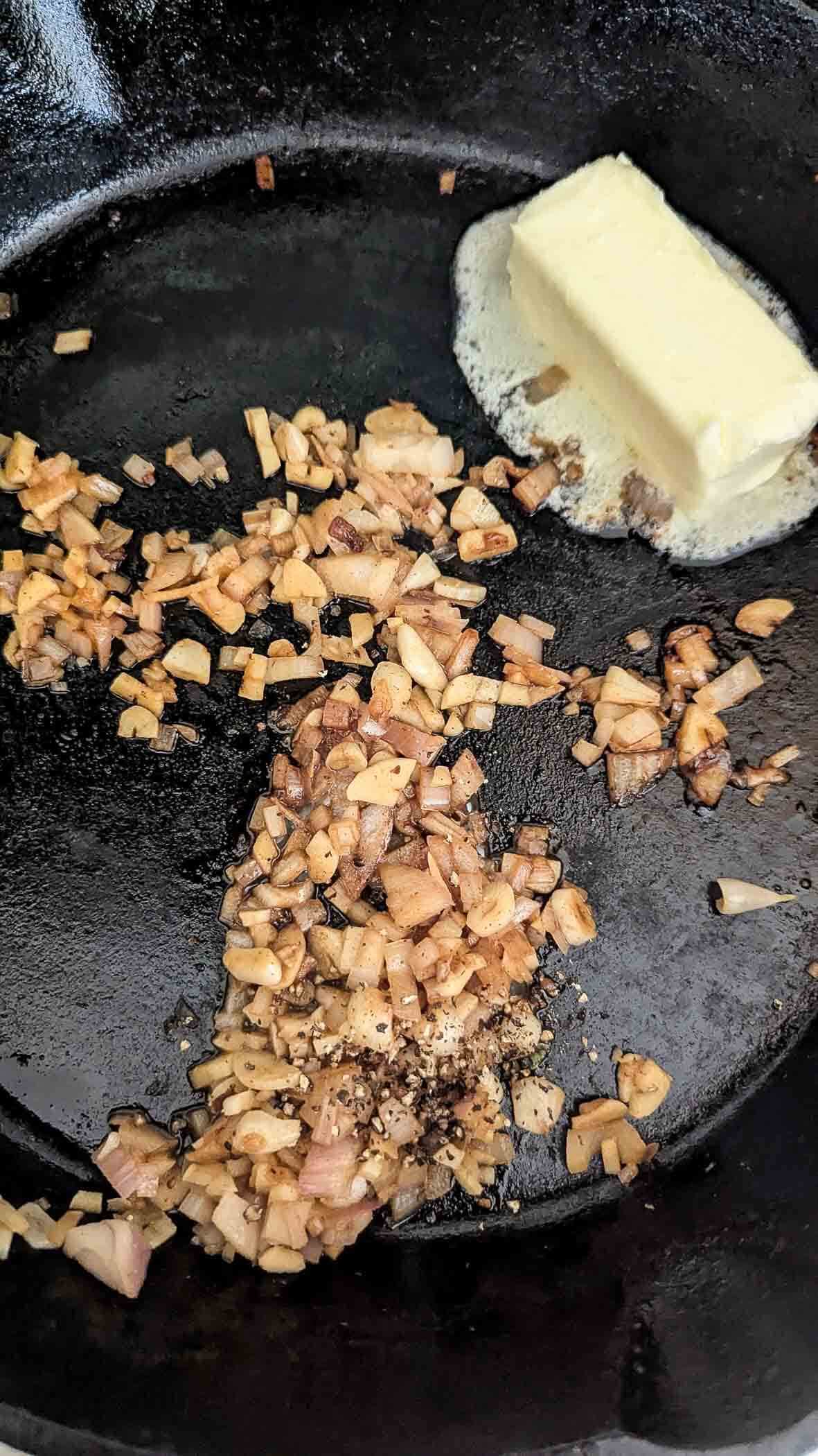 Chopped onions cooking in a black cast iron skillet, with a pat of butter melting nearby. The onions are partially browned and appear to be sautéing.