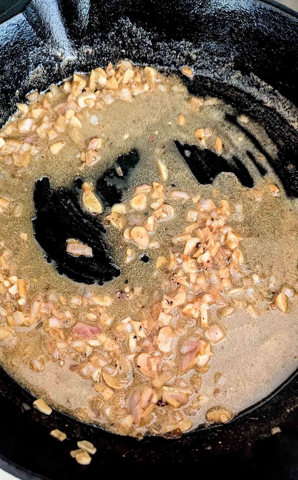Chopped garlic and shallots are being sautéed in oil in a black cast iron skillet, with some browning and bubbling visible as they cook.
