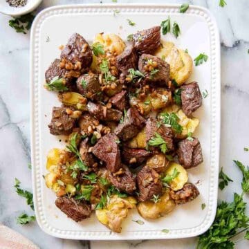 A white plate with garlic butter steak bites and smashed potatoes, garnished with chopped parsley. The dish is arranged neatly and photographed from above on a marble countertop.