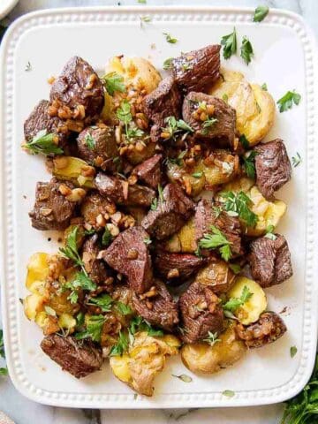 A white plate with garlic butter steak bites and smashed potatoes, garnished with chopped parsley. The dish is arranged neatly and photographed from above on a marble countertop.