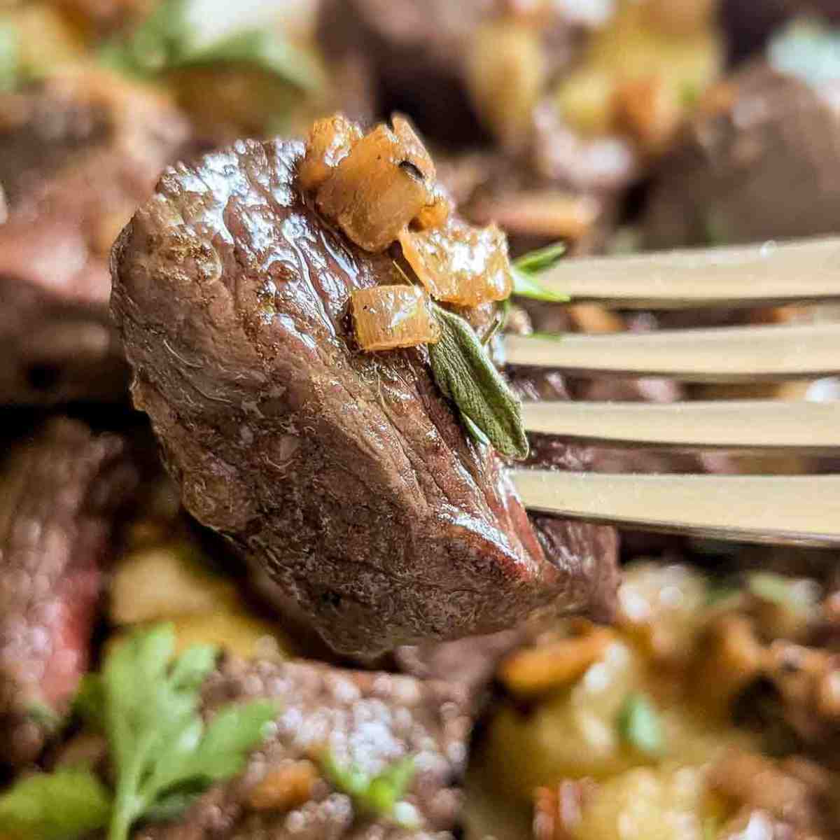 A close-up of a fork holding a juicy, cooked piece of steak topped with sautéed onions and garnished with a fresh herb. The background shows more steak and garnish out of focus.