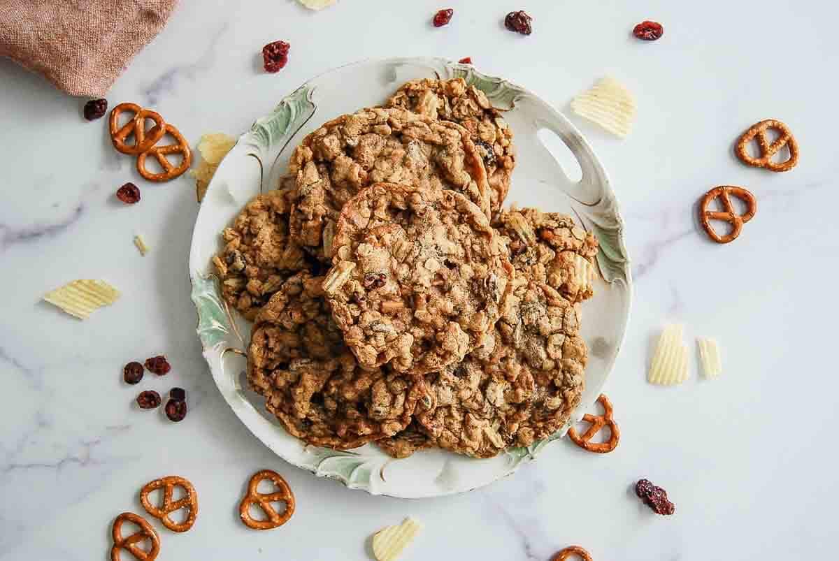 A plate of oatmeal cookies sits on a white marble surface, surrounded by small pretzels, potato chips, and dried cranberries. Some cookies and snacks are scattered around the plate.