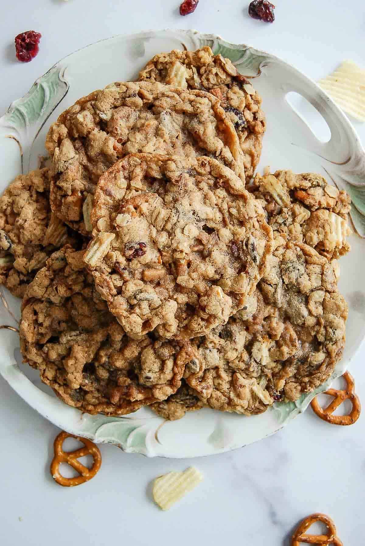 A plate of homemade cookies with visible oats, raisins, and bits of chips, surrounded by pretzels, potato chips, and dried cranberries on a white surface.