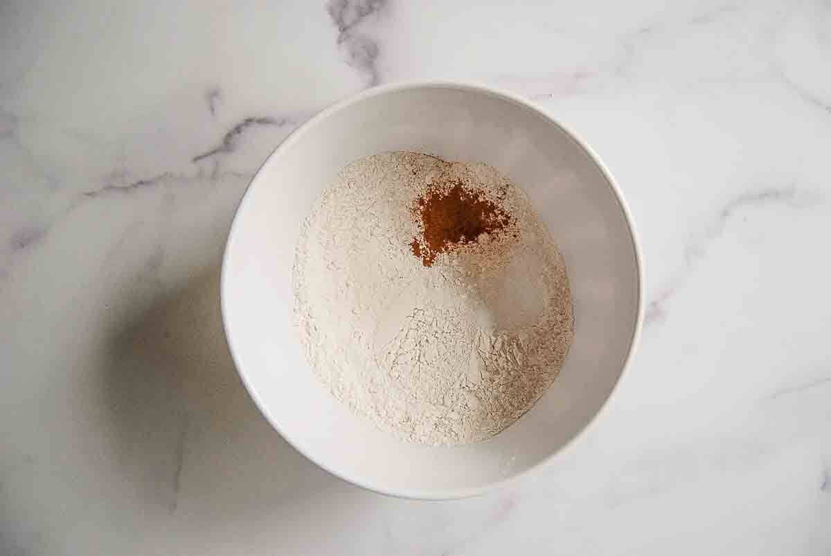 A white bowl on a marble surface containing flour and a small amount of brown spice, likely cinnamon, placed on top.