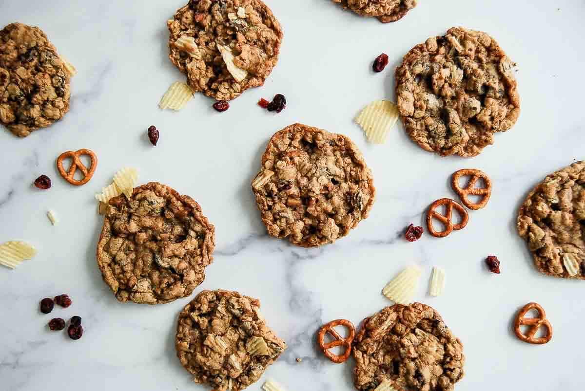 Oatmeal cookies are scattered on a white marble surface alongside mini pretzels, potato chips, and dried cranberries.