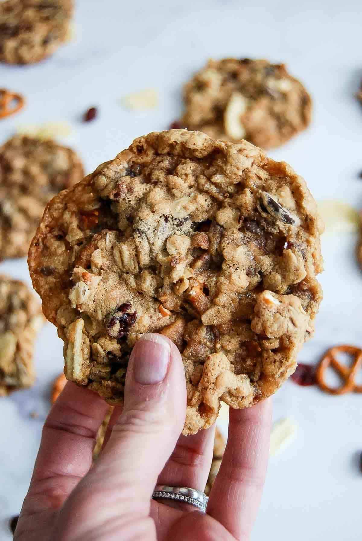A hand holds a large oatmeal cookie filled with visible pretzel pieces, raisins, and nuts. More cookies and pretzel pieces are scattered on a white surface in the background.