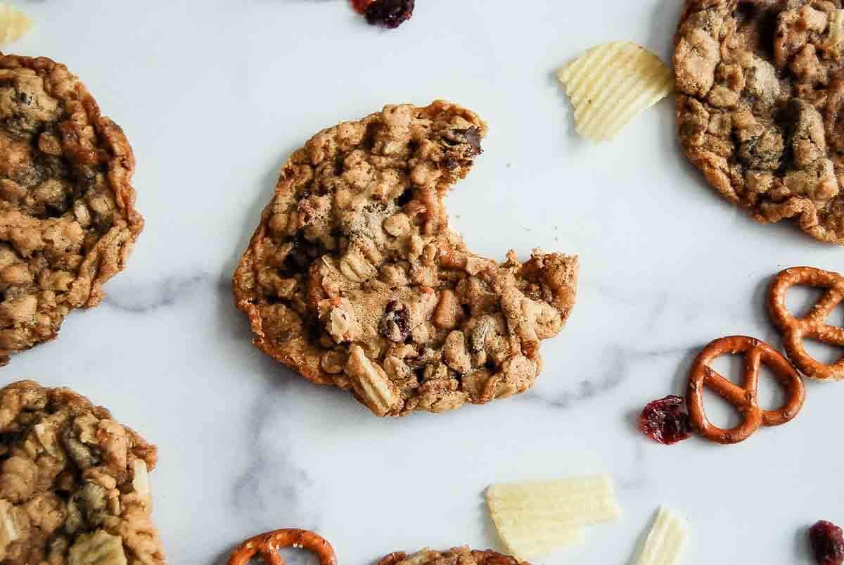 A close-up of an oatmeal cookie with a bite taken out, surrounded by potato chips, pretzels, and other cookies on a white surface.
