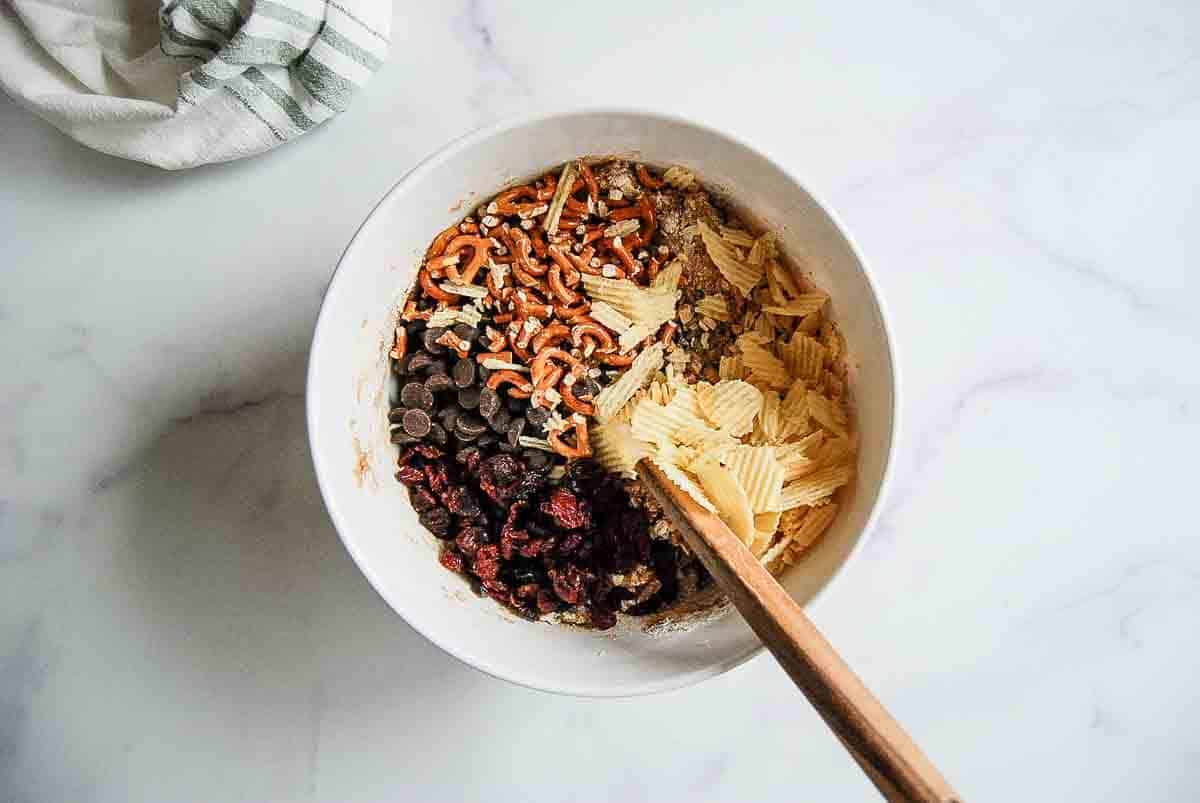 A white bowl with chocolate chips, pretzel sticks, dried cranberries, ridged potato chips, and chopped nuts, all unmixed, with a wooden spoon resting inside. A striped cloth is partially visible in the top left corner.