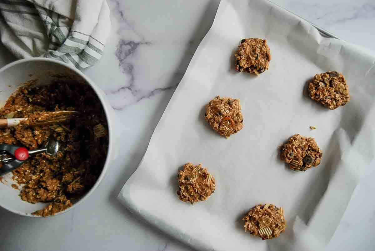 Six unbaked oatmeal cookies on a parchment-lined baking sheet, next to a mixing bowl filled with cookie dough and a red-handled scoop on a marble countertop.