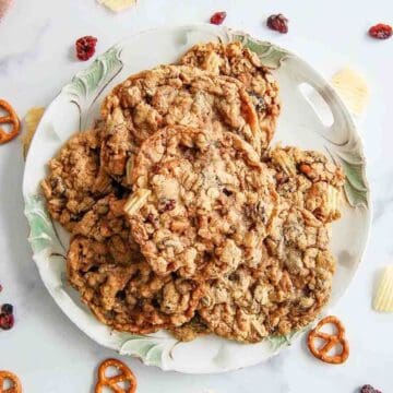 A plate filled with kitchen sink cookies sits on a white surface, surrounded by a few pretzels, potato chips, and dried cranberries.
