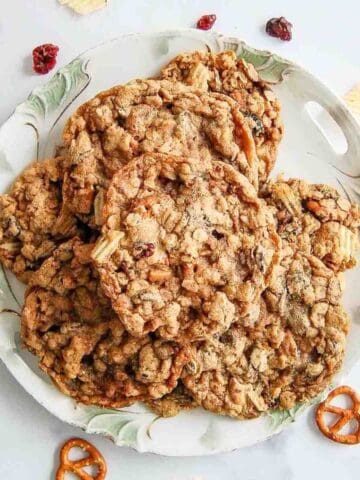 A decorative plate filled with oatmeal cookies sits on a white surface, surrounded by a few pretzels, potato chips, and dried cranberries. The cookies appear chewy with visible oats and mix-ins.