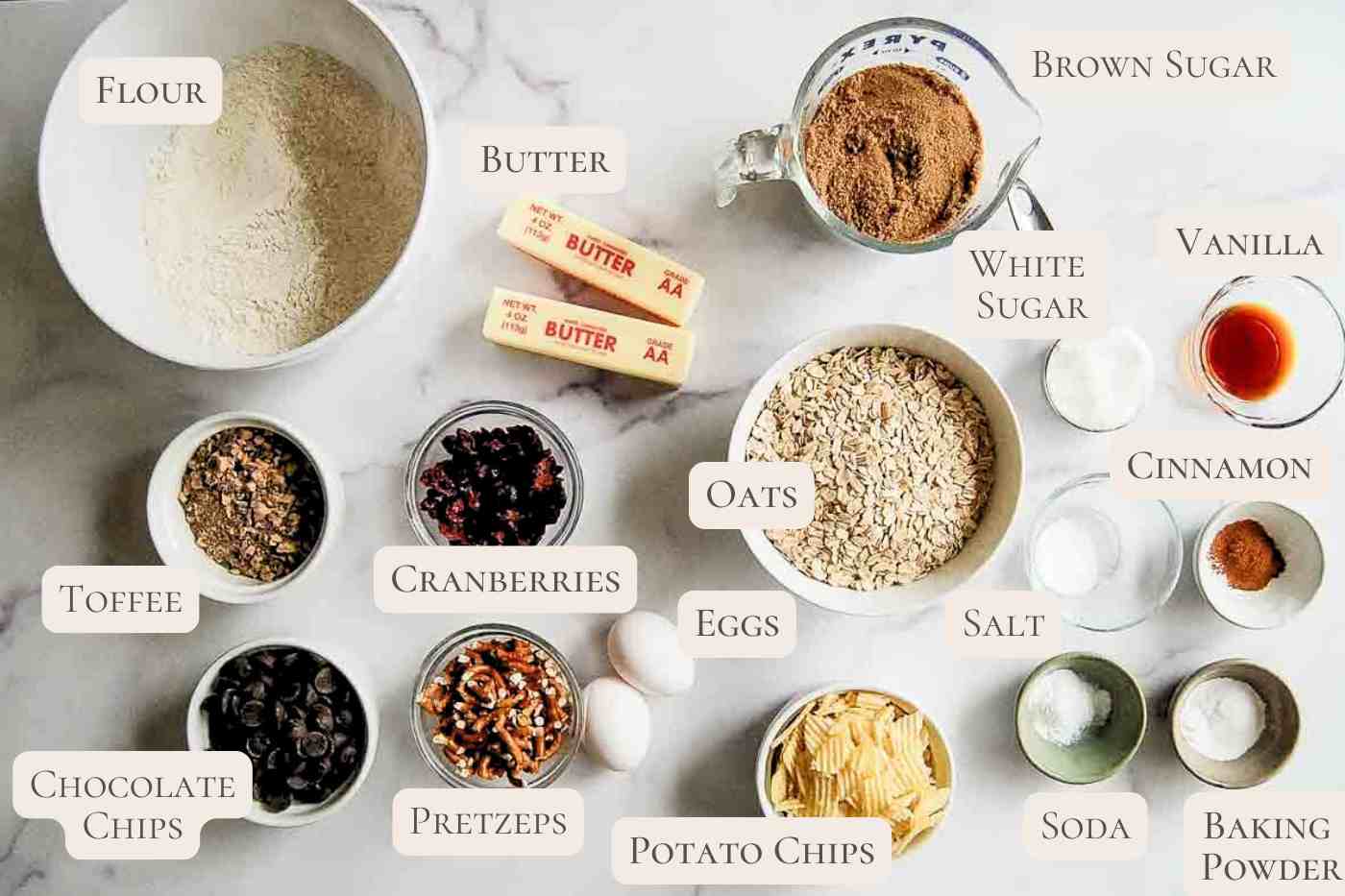 A variety of labeled ingredients for baking are spread out on a white surface, including flour, butter, brown and white sugar, oats, eggs, toffee, cranberries, chocolate chips, pretzels, potato chips, vanilla, cinnamon, salt, soda, and baking powder.