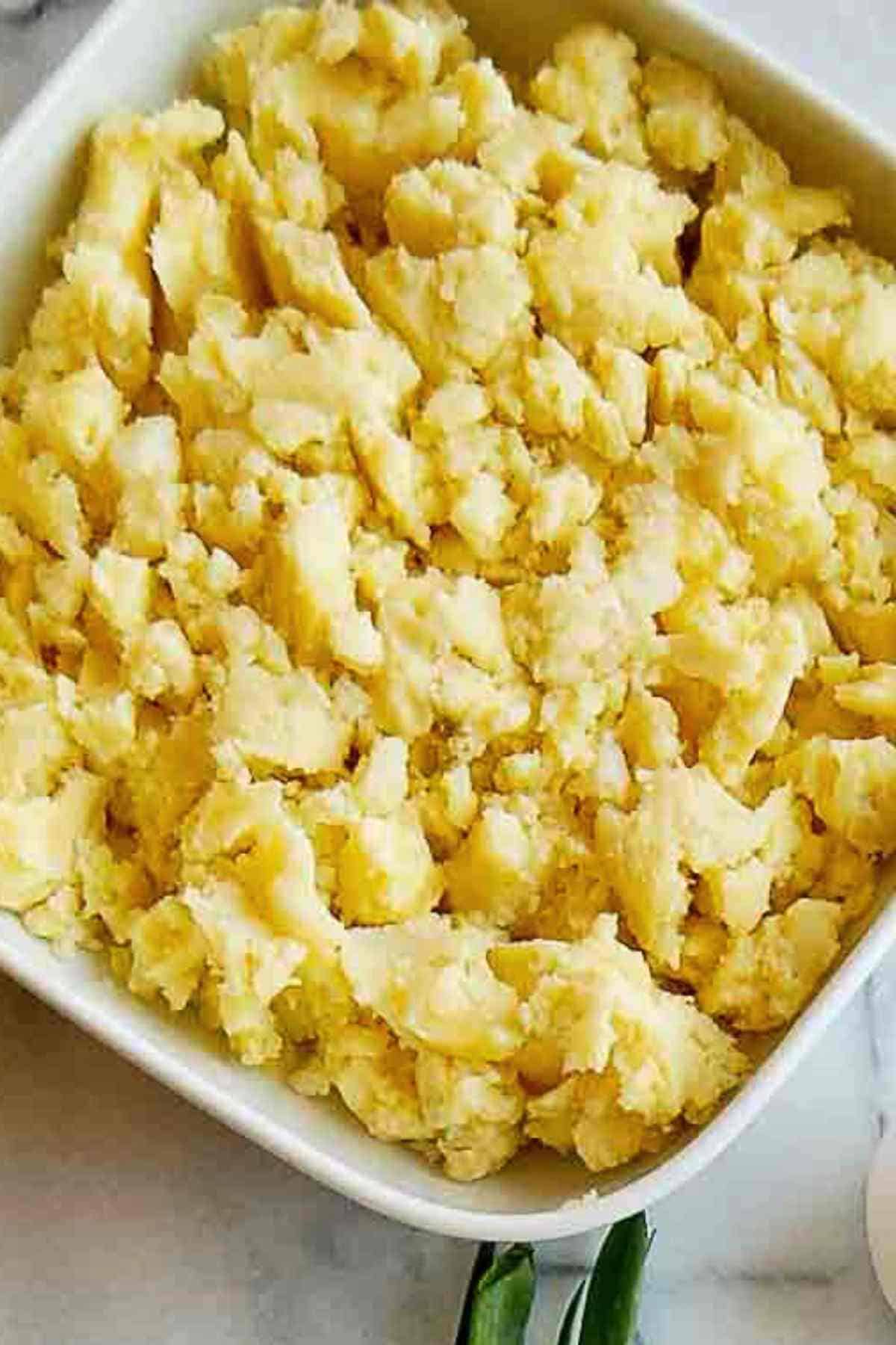 A square white dish filled with cold chunky, mashed yellow potatoes, with a slightly uneven texture.