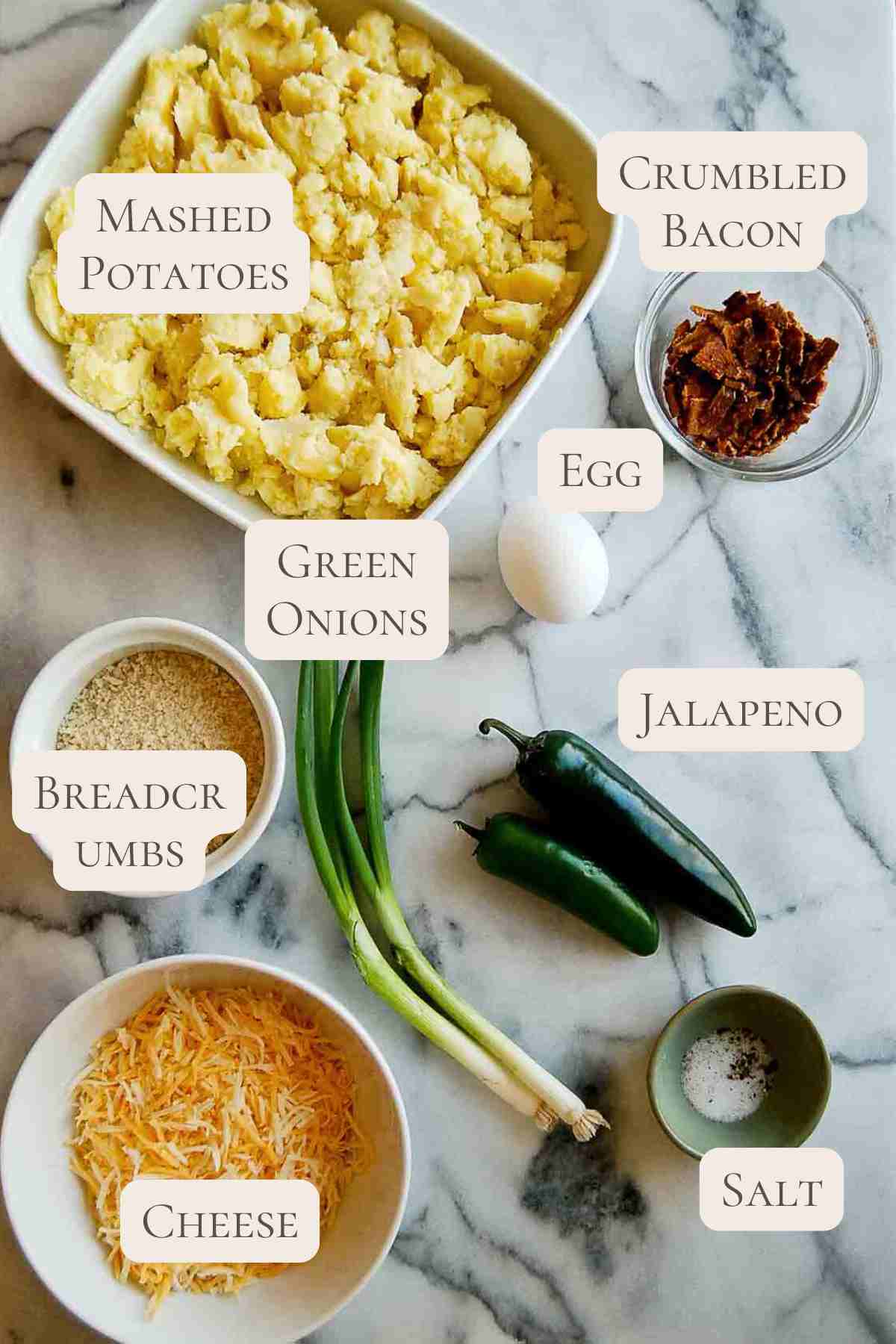A marble surface with bowls and ingredients labeled: mashed potatoes, crumbled bacon, egg, green onions, jalapeno, breadcrumbs, shredded cheese, and salt.