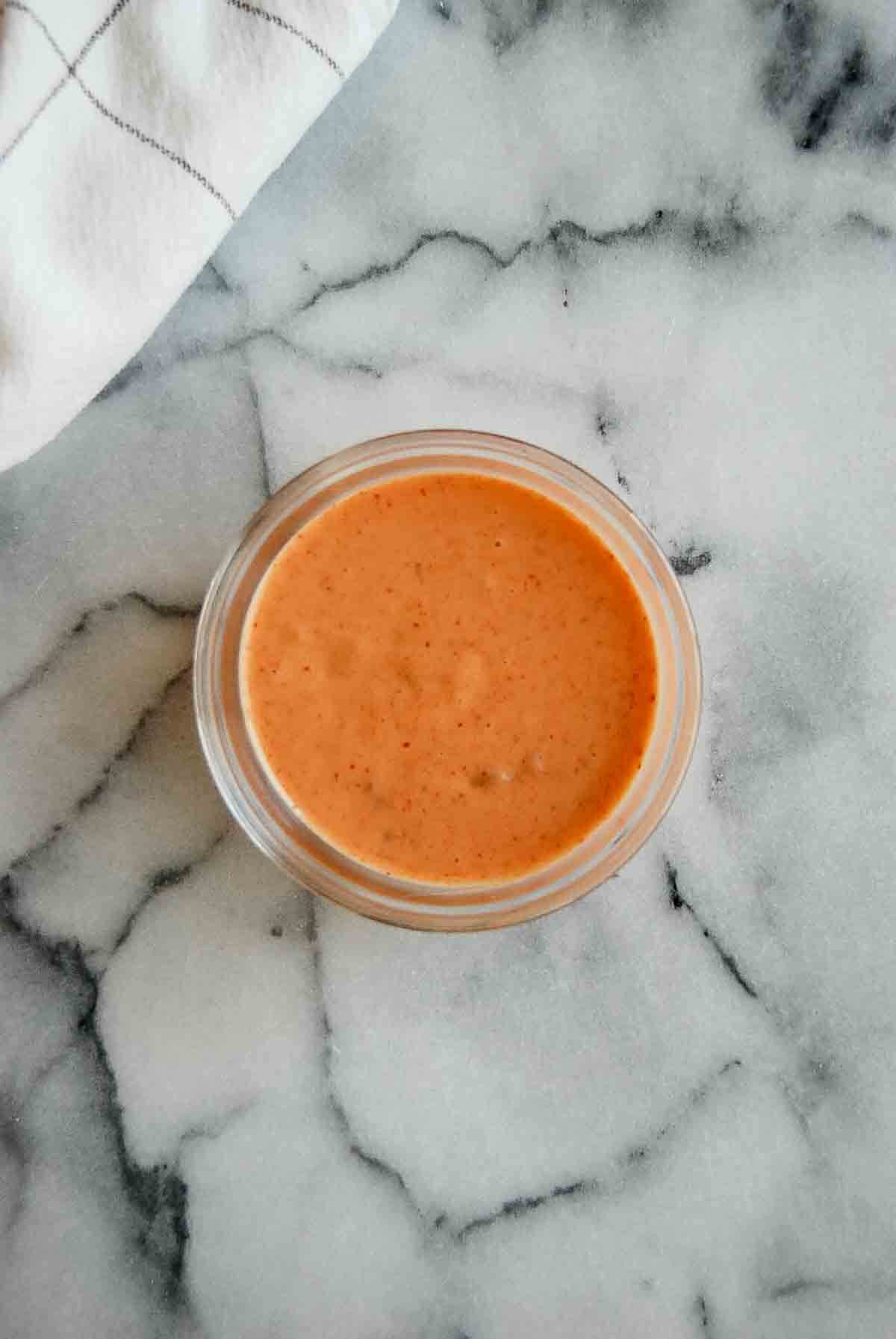 A small glass bowl filled with a creamy, orange-colored sauce sits on a marble surface next to a white cloth with gray lines.