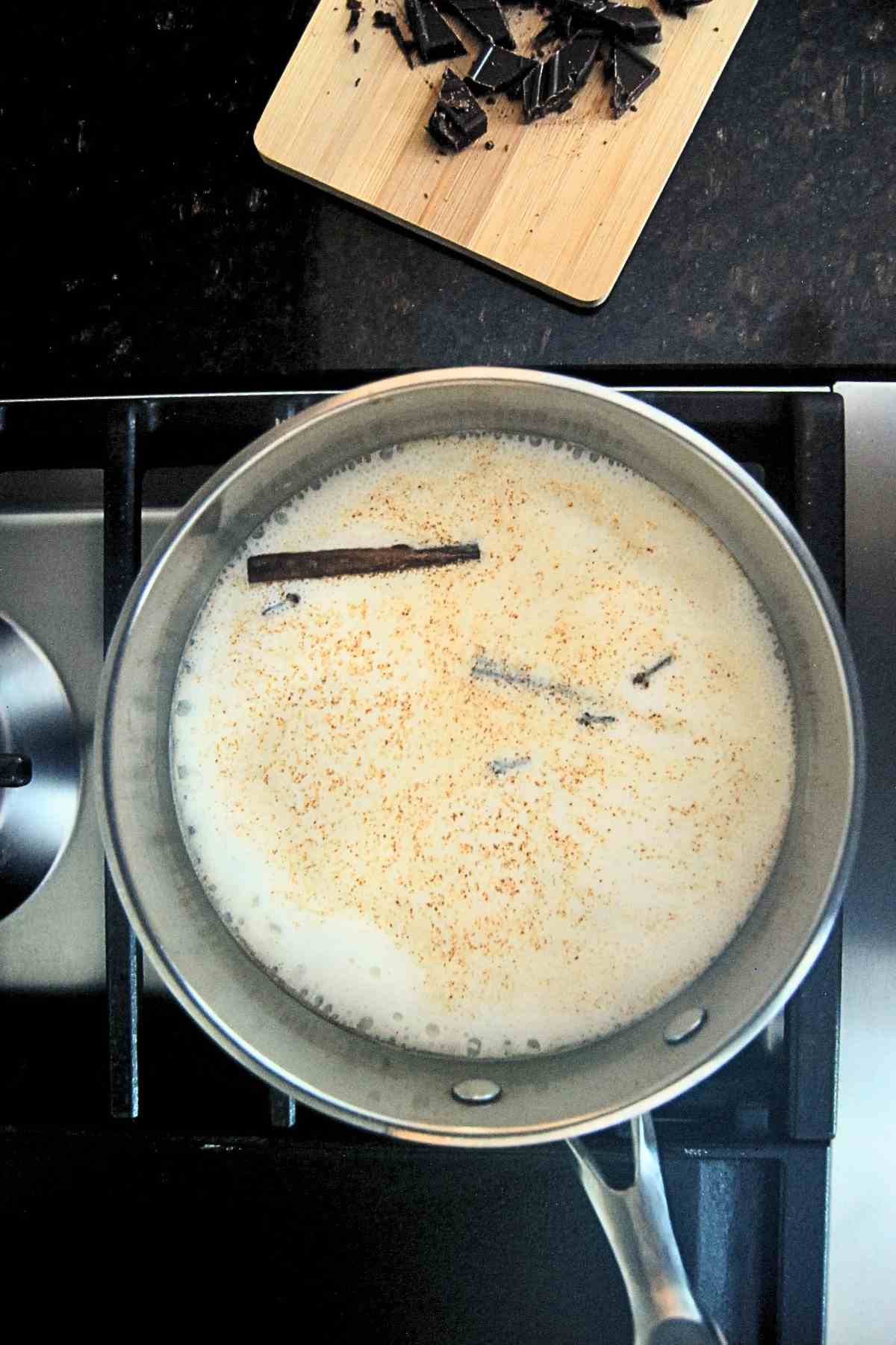 A saucepan filled with milk, cinnamon sticks, and spices heats on a stove, while chopped chocolate sits on a small wooden cutting board nearby.