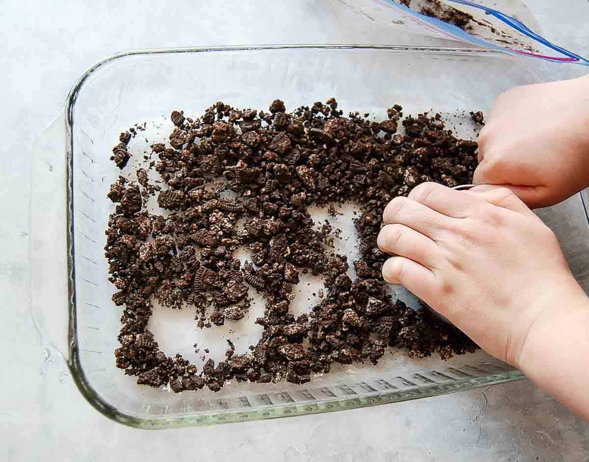 A pair of hands spreads and presses crumbled chocolate cookies in a glass baking dish on a light-colored surface.
