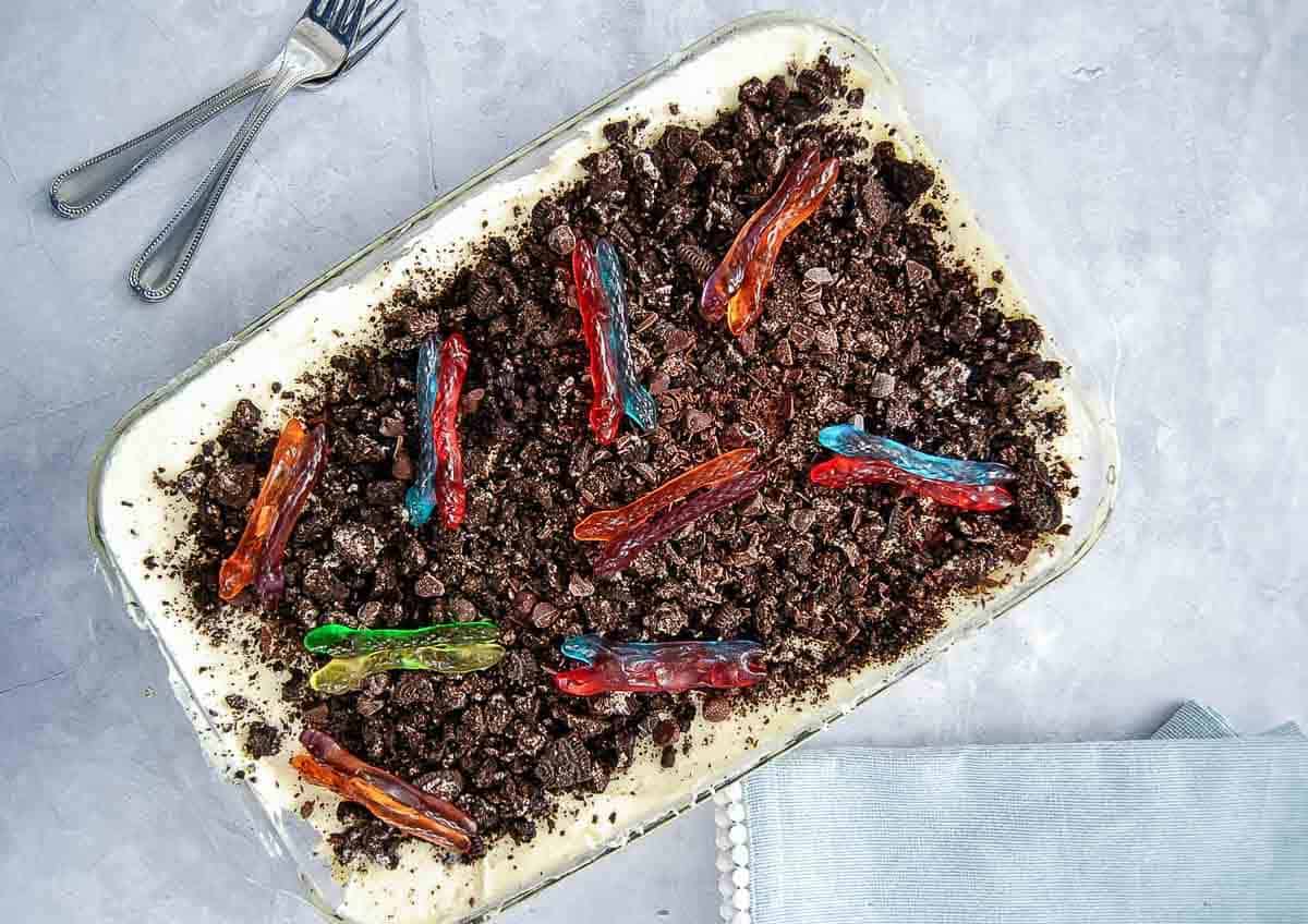 A glass dish filled with a layered dirt cake dessert topped with crumbled chocolate cookies and colorful gummy worms.