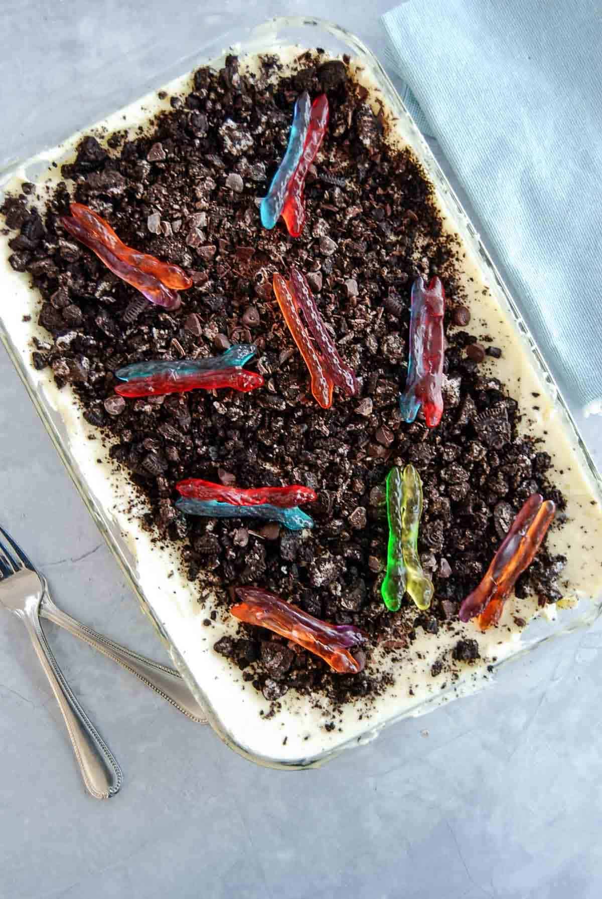 A rectangular dessert dish filled with crumbled chocolate oreo cookies on top of a creamy vanilla pudding layer, decorated with colorful gummy worms.