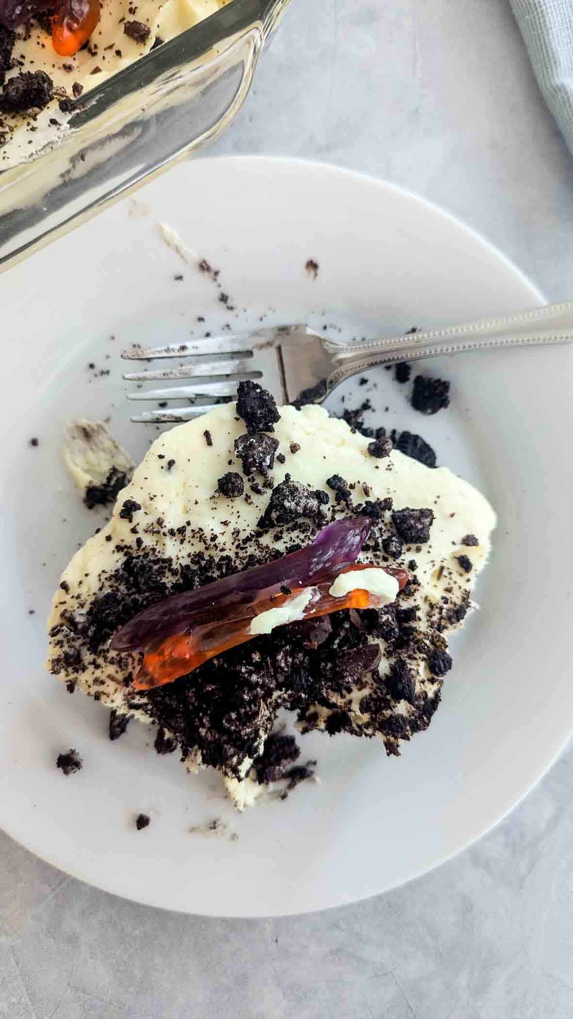 A white plate with a serving of dirt cake topped with crushed chocolate oreos and a gummy worm, next to a silver fork. Part of the dessert dish is visible at the top.