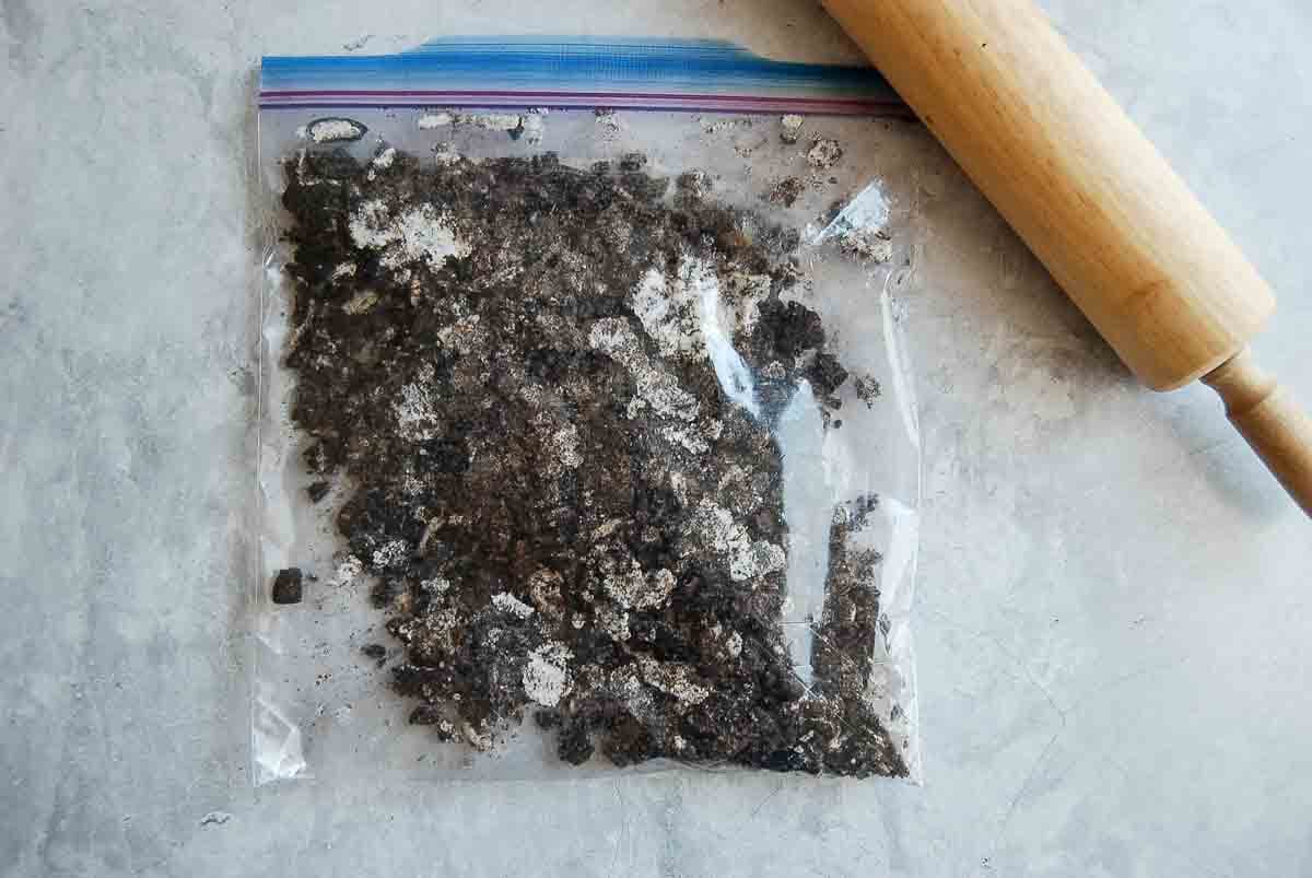 A plastic zip-top bag filled with crushed chocolate oreo cookies sits on a light gray surface next to a wooden rolling pin.