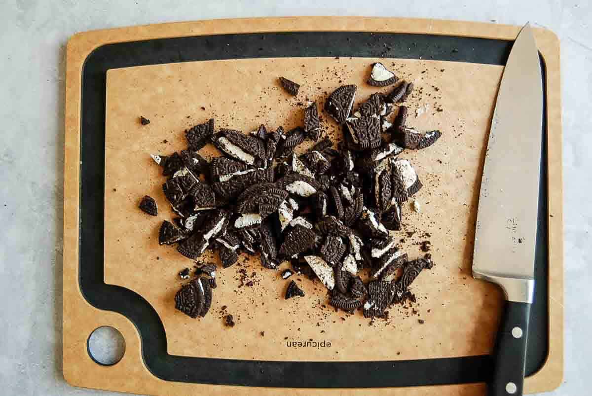A pile of roughly chopped chocolate oreo cookies with white cream filling on a brown cutting board, next to a large kitchen knife.