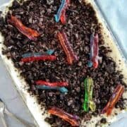 A glass dish filled with a dirt cake topped with crushed chocolate cookies and colorful gummy worms.