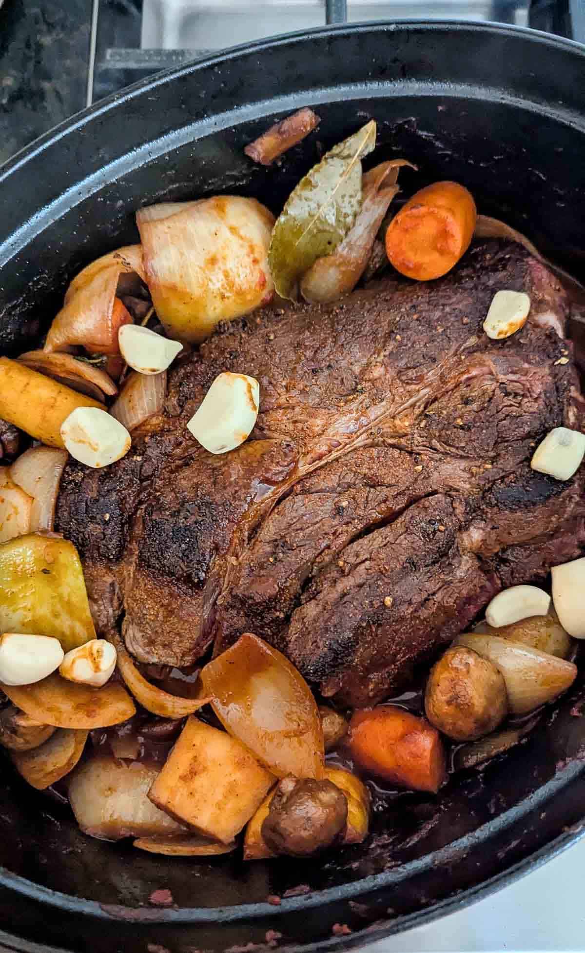 A cooked pot roast sits in a dutch oven surrounded by carrots, onions, mushrooms, and potato chunks.