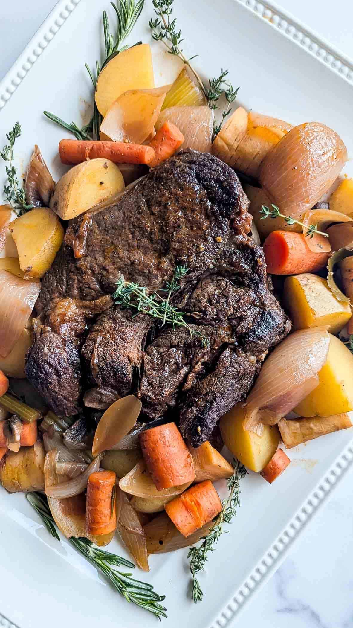 A cooked pot roast garnished with fresh herbs, surrounded by chunks of carrots, potatoes, and onions.