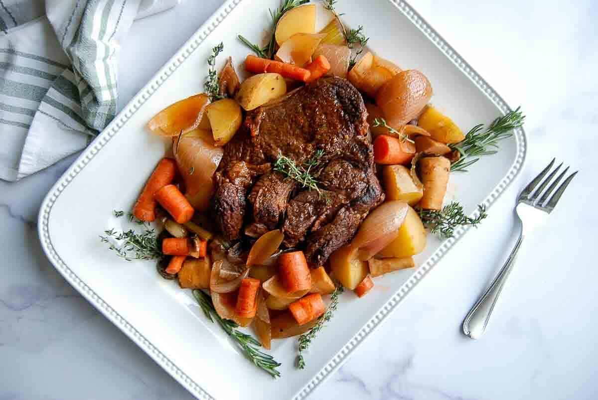 A cooked pot roast sits on a white platter, surrounded by carrots, potatoes, onions, and fresh herbs.